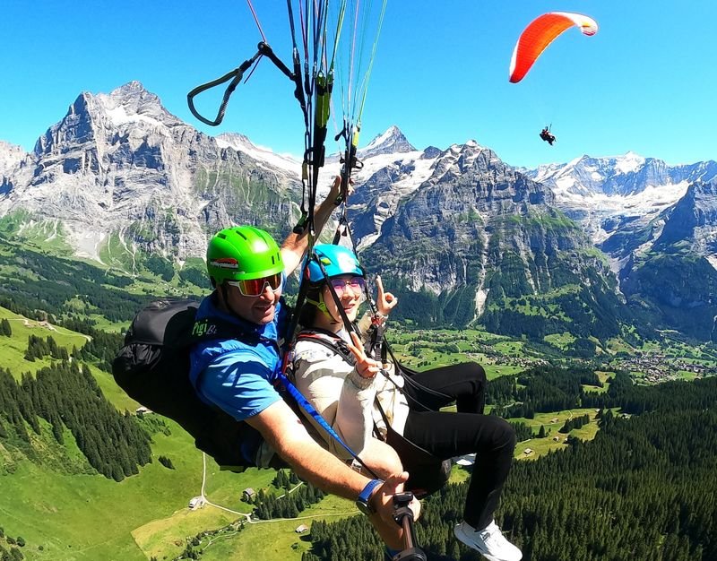 Zwei Personen beim Tandem-Paragliding mit Helmen und Gurten über einer alpinen Landschaft in der Schweiz.