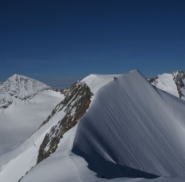 Climbers on a snowy mountain ridge with visible peaks in the background under a clear blue sky.