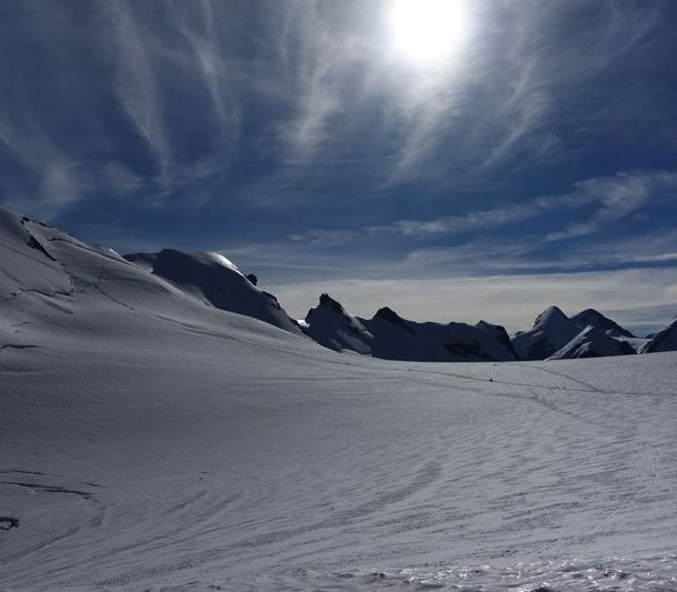 Snow-covered mountain landscape under a bright sun, with distant peaks and ski tracks visible.