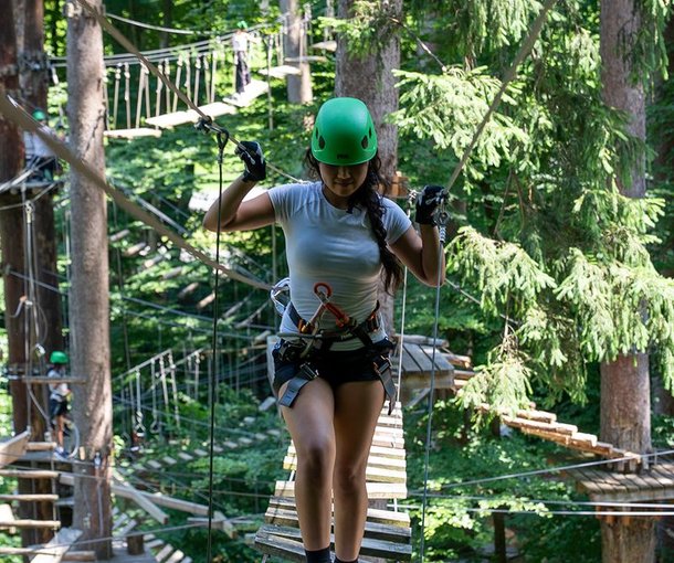 Person with a green helmet and climbing harness on a suspension bridge in the forest.