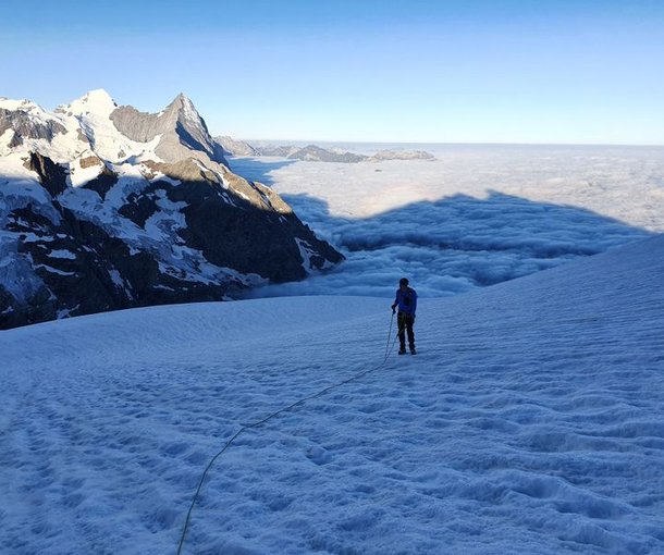 Climber on snowy mountain slope with rope and helmet, surrounded by peaks and clouds in the distance.