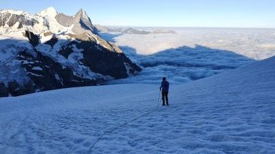 Climber on snowy mountain slope with rope and helmet, surrounded by peaks and clouds in the distance.