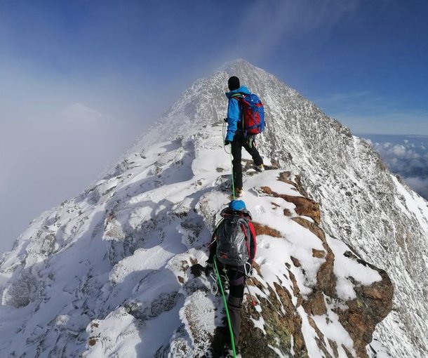 Climbers with helmets and harnesses traverse a snowy mountain ridge, roped together, under a clear sky.