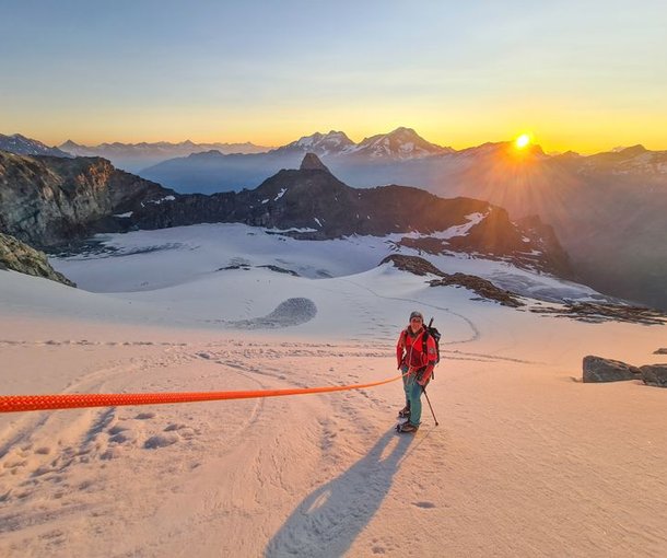 Climber in snow with helmet, harness, and rope on a mountain at sunrise.