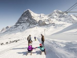 People are sledding in the snow in front of a mountain backdrop, wearing winter clothing and helmets.