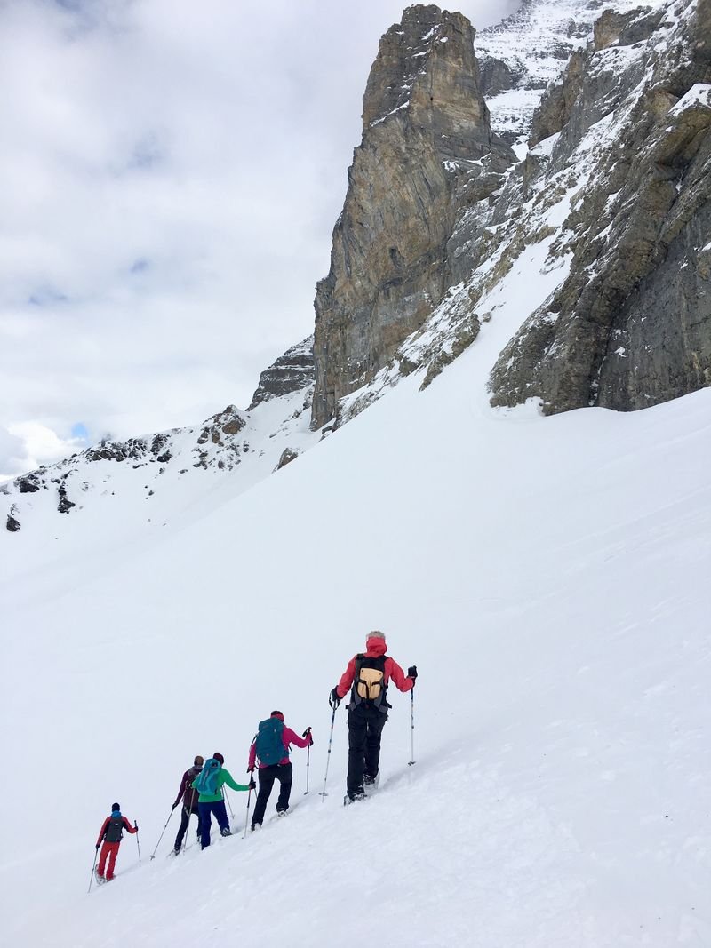 A group of people with backpacks and poles hike up a snowy mountain slope near a rocky peak.