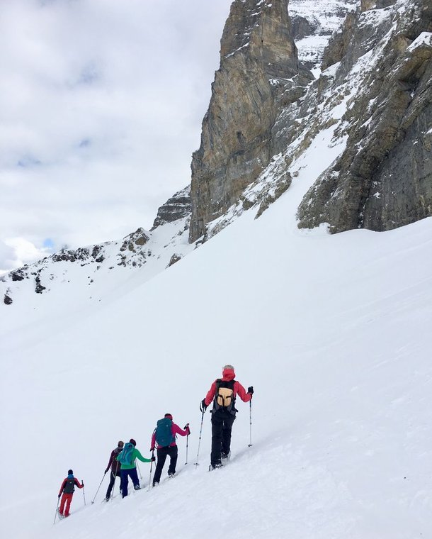 A group of people with backpacks and poles hike up a snowy mountain slope near a rocky peak.
