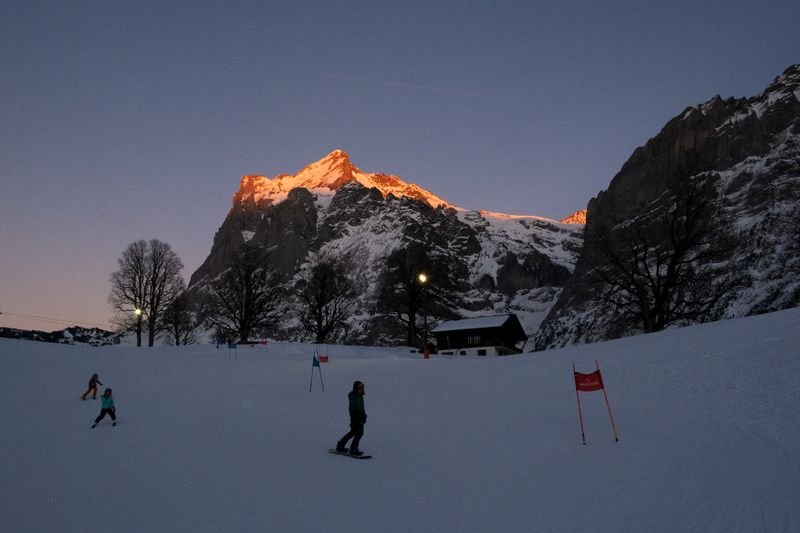 Personen fahren bei Sonnenuntergang auf einer Skipiste, umgeben von schneebedeckten Bergen.