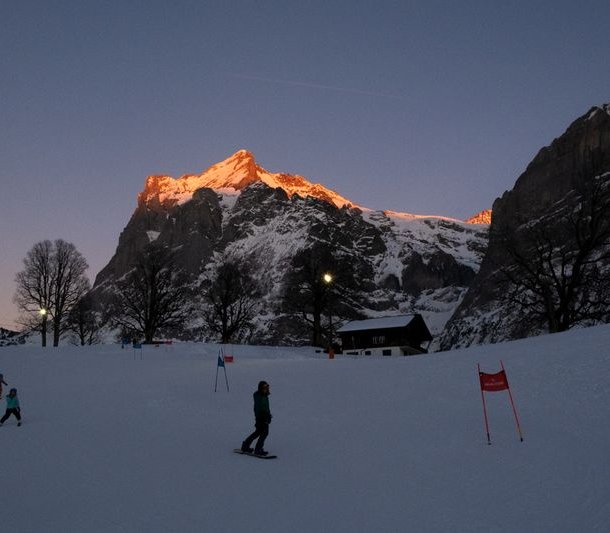 People are skiing on a slope at sunset, surrounded by snow-covered mountains.