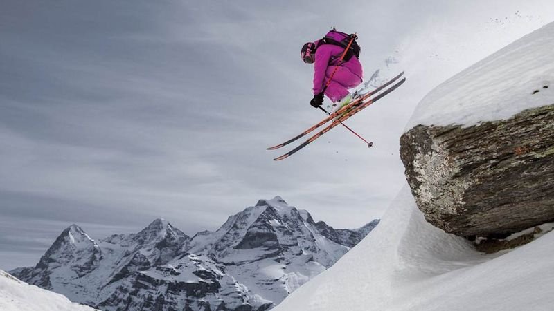 Skier in a pink suit jumps from a snowy slope, mountains in the background, wearing a helmet and backpack.