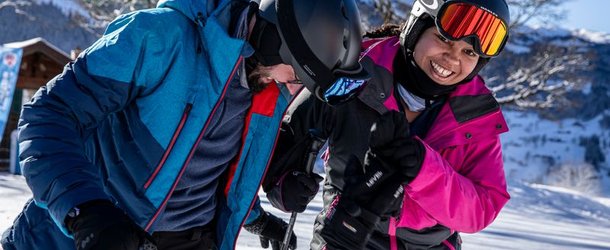 Two people with helmets and ski equipment on a snow-covered slope in the mountains.
