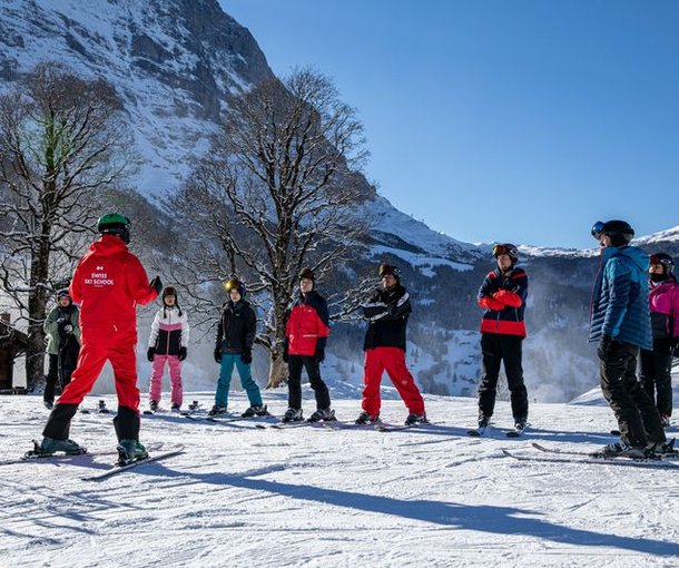 Ski instructor in red suit leads a group of skiers wearing helmets and goggles on a snowy mountain slope.