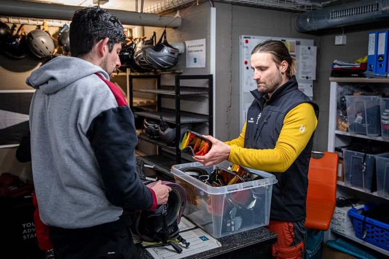 Two people in a gear room, one handing ski goggles to the other, surrounded by helmets.