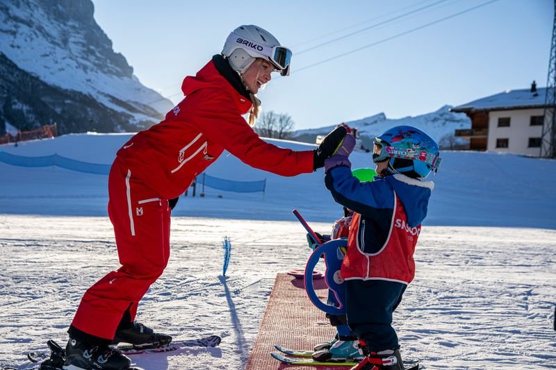 Ski instructor in red gear gives instructions to a child wearing a helmet and skis on a snow-covered slope.