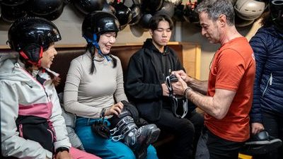 Menschen in Helmen sitzen drinnen und bereiten sich auf eine Aktivität vor, wobei ein Guide bei der Ausrüstung hilft.