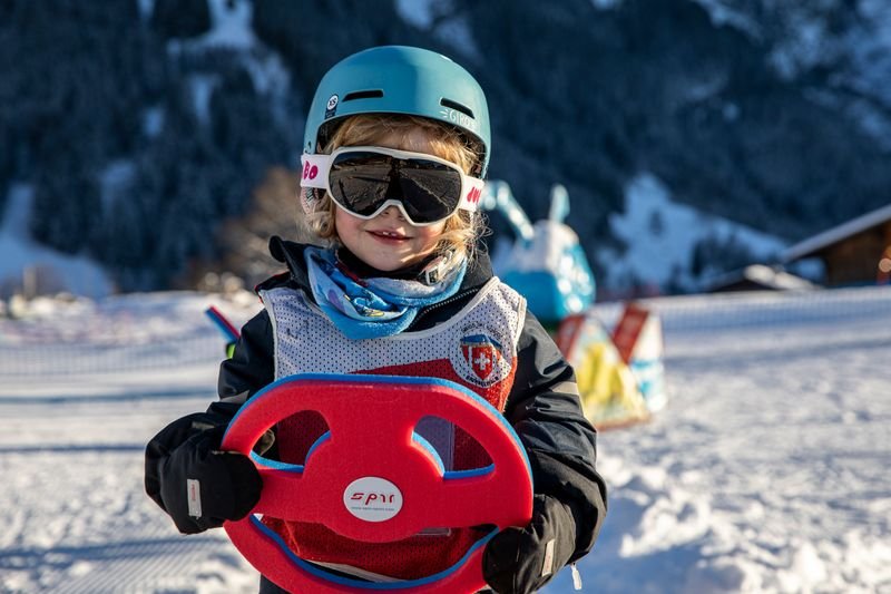 Child on a snowy slope with helmet, ski goggles, and steering wheel toy, mountains in the background.