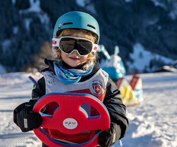 Child on a snowy slope with helmet, ski goggles, and steering wheel toy, mountains in the background.