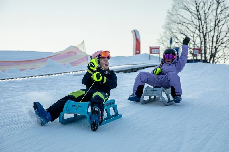 Zwei Personen rodeln auf Schnee, tragen Helme und Skibrillen. Hintergrund: verschneite Landschaft.
