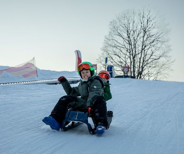 Two people are sledding on a snow-covered slope, wearing helmets and winter clothing.