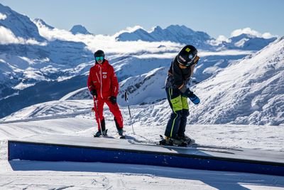 Personen mit Skiern auf einer Piste in den Bergen; eine Person trägt rote Kleidung, die andere schwarz.
