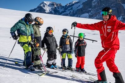 Skigruppe mit Helmen und Skistöcken auf verschneitem Berg, begleitet von einem Skilehrer in Rot.