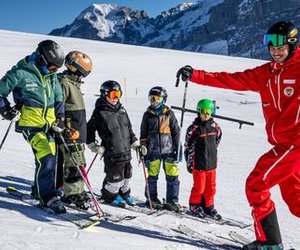 Ski group with helmets and ski poles on a snowy mountain, accompanied by a ski instructor in red.