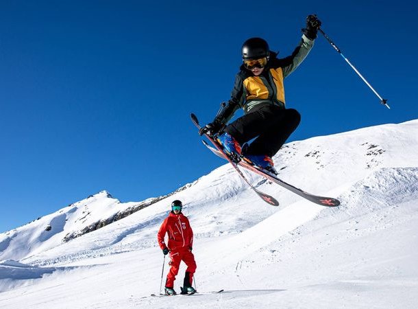 Personen beim Skifahren auf schneebedecktem Berg; eine Person springt, beide mit Helmen und Skiern.