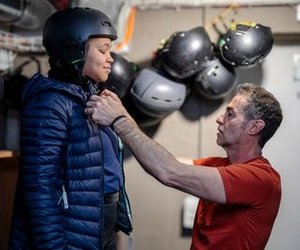 Person in helmet and jacket being assisted by another in a room with helmets on the wall.