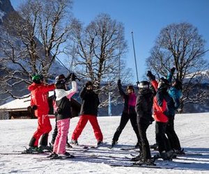Skiers in helmets and gear raise poles in a snowy mountain setting with trees and a chalet.