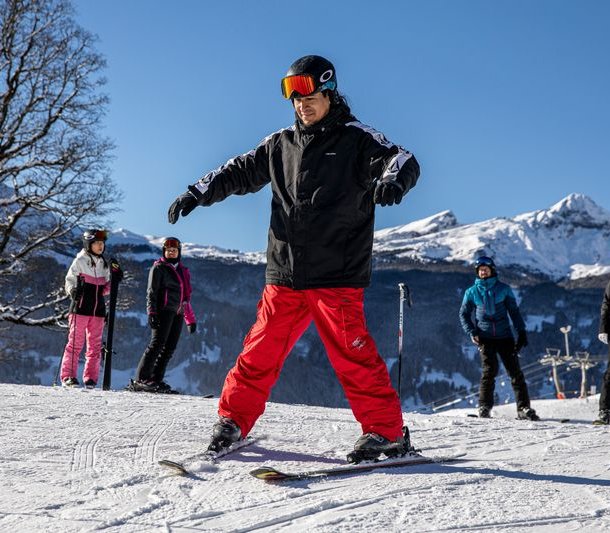 A group of skiers on a snowy mountain slope, wearing helmets and ski gear, with mountains in the background.