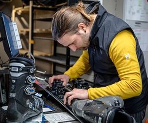 Person adjusting ski bindings indoors, with ski boots and computer on the counter.