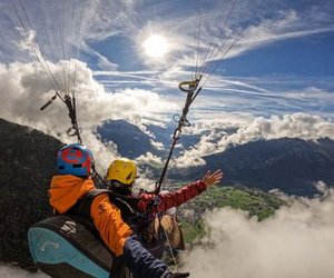 Zwei Personen beim Paragliding über einer Berglandschaft, mit Helmen und Gurten ausgestattet.