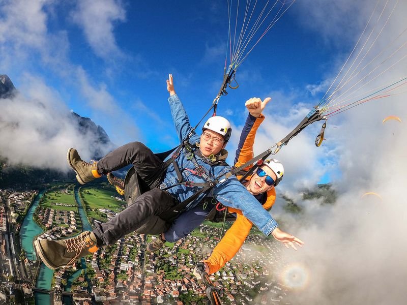 Zwei Personen beim Tandem-Paragliding über einer Stadt, mit Helmen und Gurten, umgeben von Bergen.