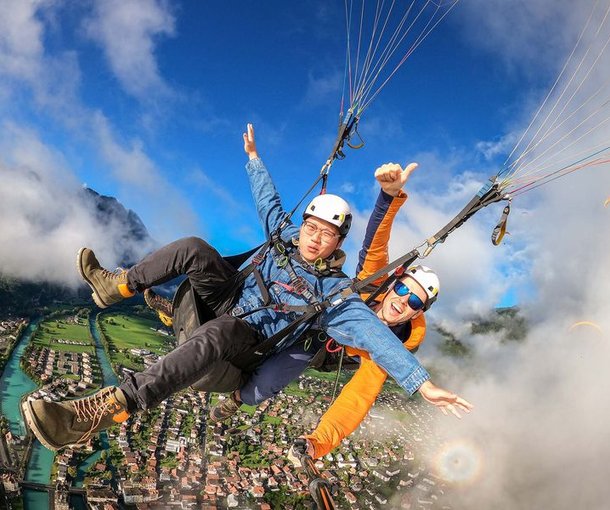 Zwei Personen beim Tandem-Paragliding über einer Stadt, mit Helmen und Gurten, umgeben von Bergen.