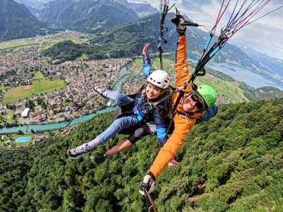 Zwei Personen beim Tandem-Paragliding über einer Stadt, mit Helmen und Gurten, umgeben von Bergen.