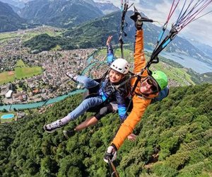 Zwei Personen beim Tandem-Paragliding über einer Stadt, mit Helmen und Gurten, umgeben von Bergen.