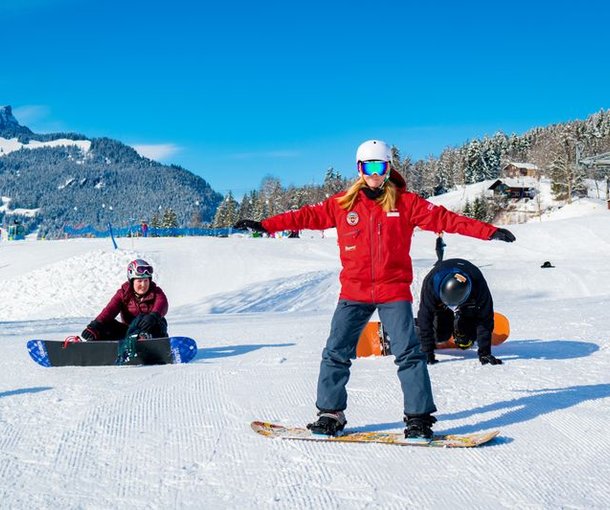 People snowboarding on a snowy slope; wearing helmets and snow clothing; mountains in the background.