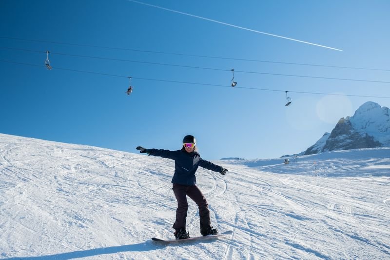 Snowboarder with helmet and ski goggles rides on a snow-covered slope, cable car in the background.
