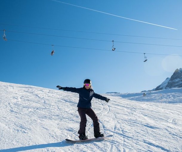 Snowboarder with helmet and ski goggles rides on a snow-covered slope, cable car in the background.