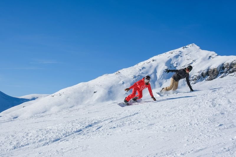 Zwei Personen snowboarden auf einer schneebedeckten Piste in den Bergen, mit Helmen und Winterkleidung.