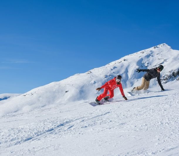 Two people are snowboarding on a snow-covered slope in the mountains, wearing helmets and winter clothing.
