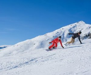 Two people are snowboarding on a snow-covered slope in the mountains, wearing helmets and winter clothing.