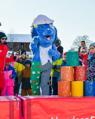 Children and adults in winter clothing are standing around a mascot on a snowy slope.