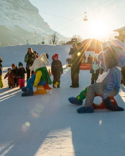 People in winter clothing and mascots on snow in front of mountain scenery and cabin at sunset.