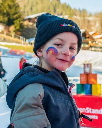 Child on ski slope with painted face, beanie, and winter jacket; snow-covered mountains in the background.