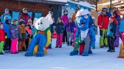 Children and adults stand on snow next to two mascots in colorful costumes in front of wooden huts.