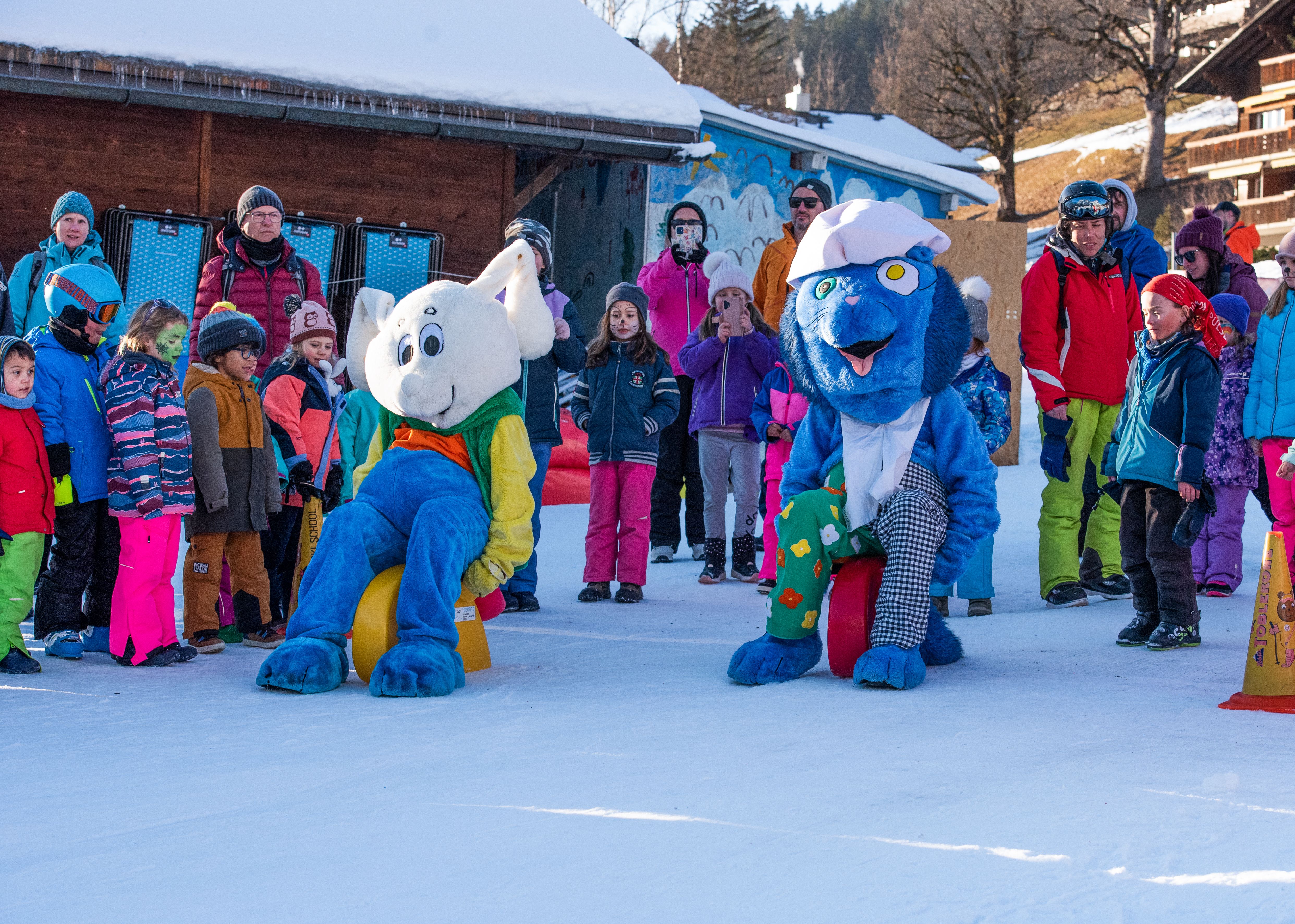Children and adults stand on snow next to two mascots in colorful costumes in front of wooden huts.