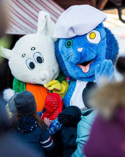 Two people in animal costumes surrounded by children in winter clothing at an outdoor event.