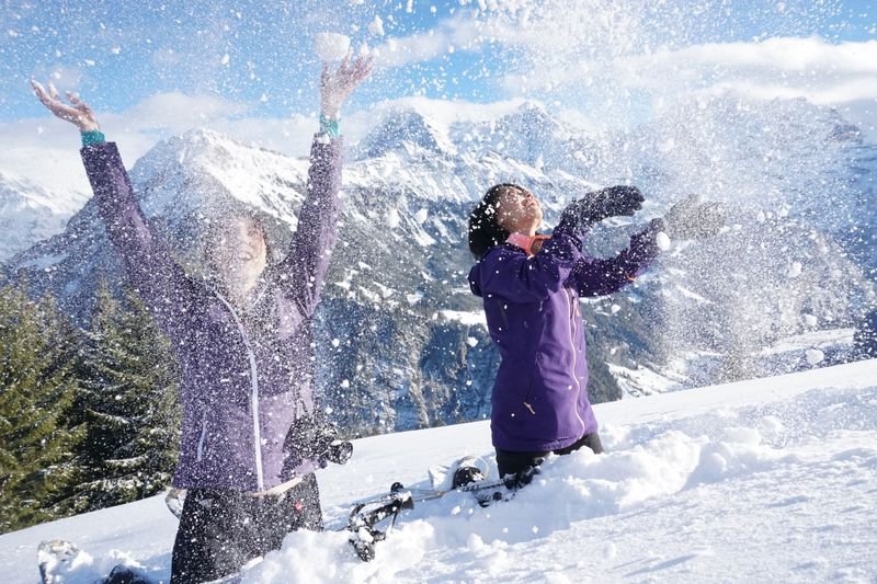 Two people are throwing snow in the air, surrounded by snow-covered mountains and trees.