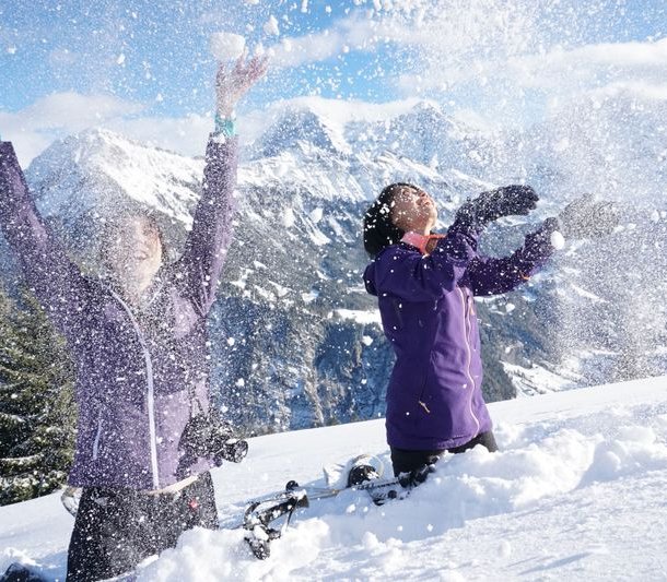 Two people are throwing snow in the air, surrounded by snow-covered mountains and trees.